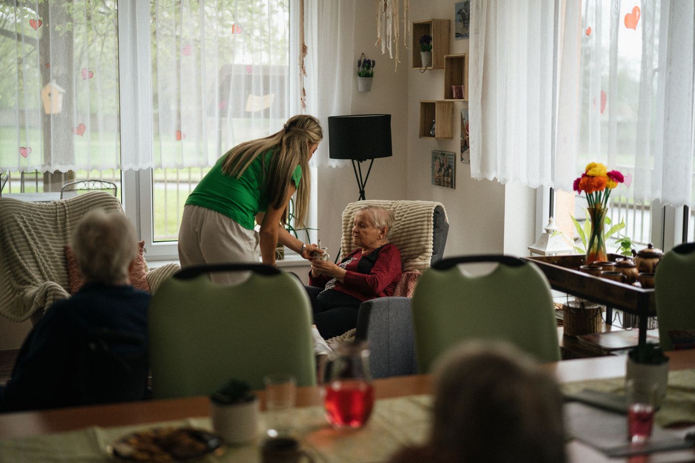 A caregiver bringing afternoon tea to a resident in a bright, plant-filled common room